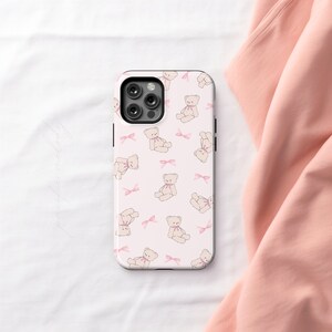 May include: A white phone case with a pink and white teddy bear pattern. The case has a cutout for the camera lens.