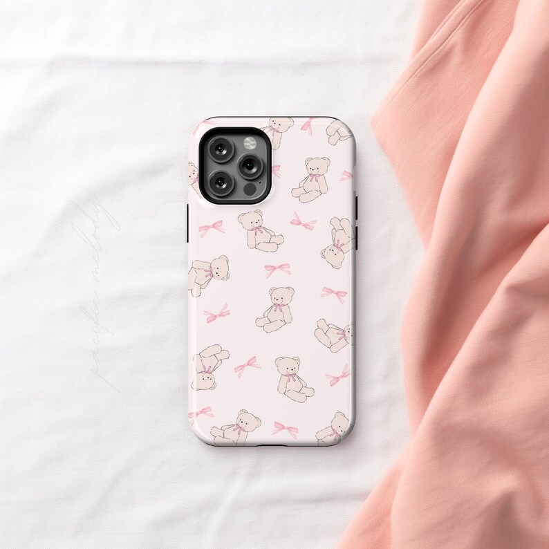 May include: A white phone case with a pink and white teddy bear pattern. The case has a cutout for the camera lens.