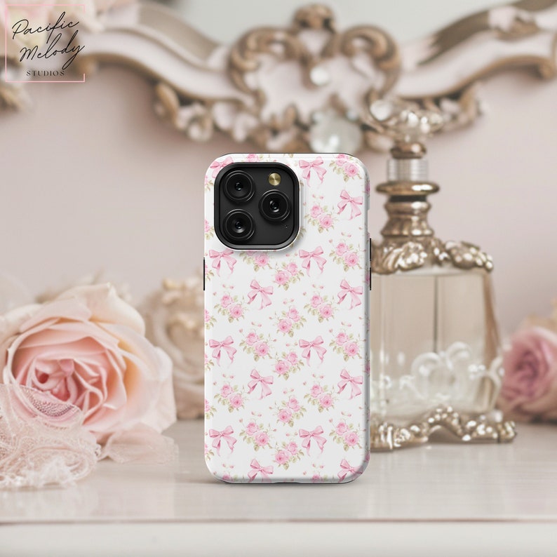 May include: A white phone case featuring a pink floral pattern and pink bows. The case is designed for a phone with a square camera module.