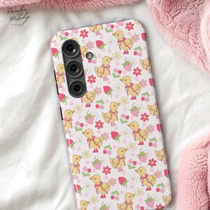 May include: A white phone case with a pink and yellow pattern of ducks, strawberries, and flowers. The case has a black camera lens.