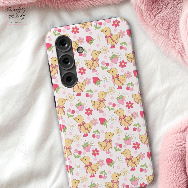 May include: A white phone case with a pink and yellow pattern of ducks, strawberries, and flowers. The case has a black camera lens.