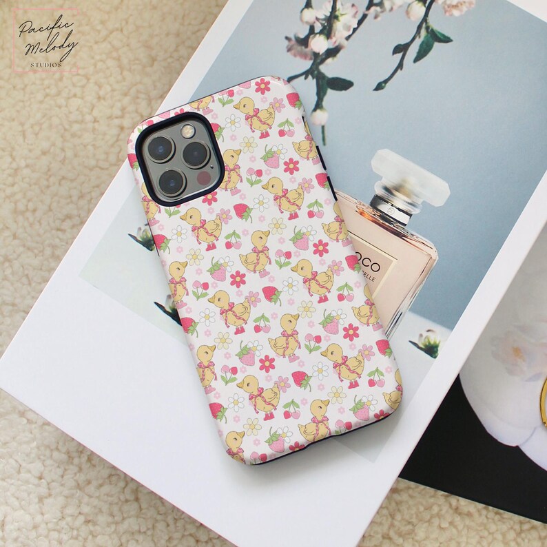 May include: A white phone case with a pink and yellow duck and strawberry pattern. The case has a black border and a camera cutout.