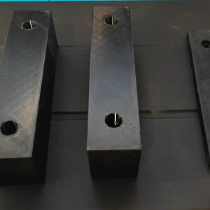 Peut inclure: Trois blocs rectangulaires noirs de tailles variables. Chaque bloc est percé d'un trou. Les deux blocs les plus grands ont un trou plus petit en haut. Les blocs sont disposés sur une surface sombre.