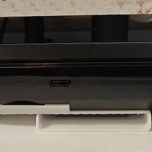 May include: A black Nintendo Switch console with a white stand. The console is closed and the screen is off.