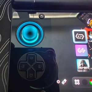 May include: A black handheld gaming console featuring a glowing cyan circular joystick, a black directional pad with four arrow buttons, and a glossy screen displaying a digital interface with various application icons and the text Call of Duty.