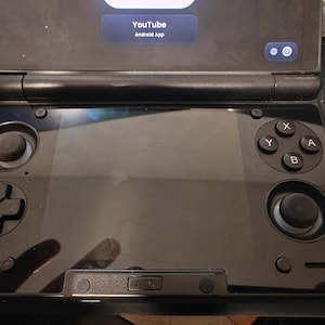 May include: A black handheld gaming console featuring a central screen, a directional pad on the left, and four action buttons labeled X, Y, A, and B on the right. Two analog sticks are present, and the screen displays the YouTube Android App.