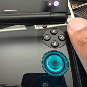 May include: A handheld gaming device with a black glossy finish, featuring a four-button cluster labeled X, Y, A, and B, and a circular joystick illuminated with a bright cyan light. The screen displays settings including Smooth Display 60.0.