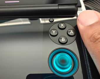 May include: A handheld gaming device with a black glossy finish, featuring a four-button cluster labeled X, Y, A, and B, and a circular joystick illuminated with a bright cyan light. The screen displays settings including Smooth Display 60.0.