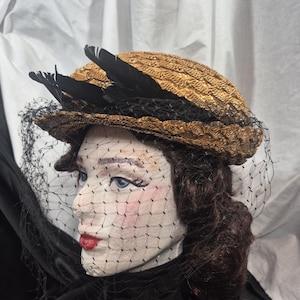 Puede incluir: Un sombrero de estilo vintage con una corona tejida de color marrón dorado, adornado con plumas negras y un velo de red negro. El sombrero está sobre una cabeza de maniquí con rasgos pintados, incluyendo ojos azules y labios rojos, creando un look clásico y elegante.