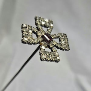 Puede incluir: Broche plateado en forma de cruz con una piedra central rectangular de color granate. La cruz está incrustada con pequeños diamantes de imitación transparentes. Una aguja larga y fina se extiende desde la parte posterior.