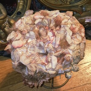 May include: A rounded hat covered in numerous layered fabric petals in shades of dusty pink, muted mauve, and faded beige. The textured surface features overlapping textile pieces that create a dense, floral-like appearance across the entire dome.