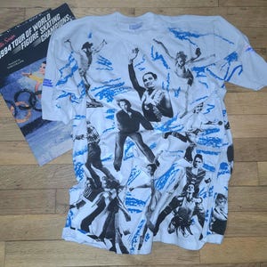 Könnte beinhalten: Weißes T-Shirt mit Schwarz-Weiß-Bildern von Eiskunstläufern und blauen Akzenten. Das Shirt hat einen Rundhalsausschnitt und kurze Ärmel. Ein Buch mit dem Titel "Campbell's Soups 1994 Tour of World Figure Skating Champions" ist sichtbar.