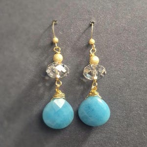 May include: A pair of gold-colored dangle earrings. Each earring features a light blue, teardrop-shaped stone, a clear faceted bead, and a gold-colored bead. The earrings are suspended from gold-colored ear wires.