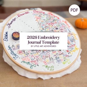Può includere: Un telaio da ricamo con tessuto bianco con ricami colorati e il testo "2026 Embroidery Journal Template". Il design include mesi, piccole illustrazioni e una piccola zucca. Il telaio è in legno.