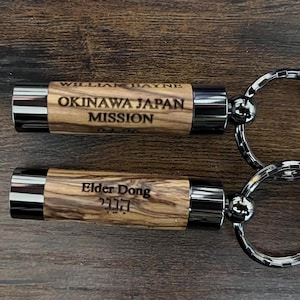 May include: Two wooden keychains with silver-colored metal accents and key rings. One keychain reads "WILLIAM BAYNE OKINAWA JAPAN MISSION" and the other reads "Elder Dong" with Hebrew text. The keychains are on a wooden surface.