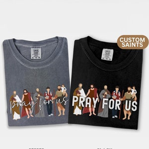 May include: Two t-shirts with a graphic of a group of people with the text "pray for us". The shirt on the left is a light gray color with the text in a cursive font. The shirt on the right is black with the text in a bold font.