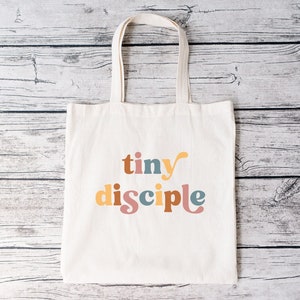 以下が含まれることがあります： カラフルなスタイリッシュな文字で「tiny disciple」と印刷された白いキャンバス地のトートバッグです。