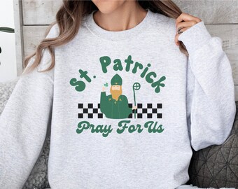 May include: A gray sweatshirt with a green and black graphic print of a cartoon St. Patrick holding a staff and a shamrock. The text "St. Patrick Pray For Us" is printed in a retro font.