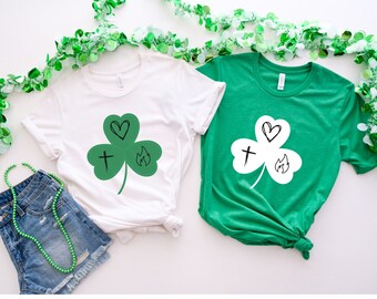 May include: Two t-shirts with a green shamrock design. The shamrock has a heart, cross, and flame inside. One shirt is white and the other is green.