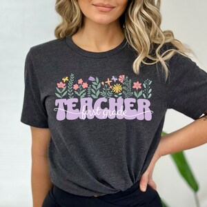 May include: A dark grey t-shirt with a purple graphic that says "TEACHER first grade" with a row of colourful flowers above the text.