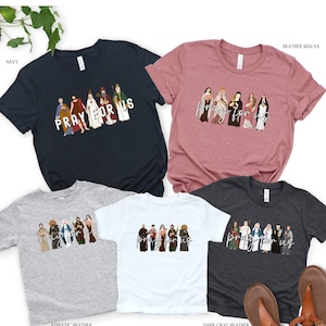 May include: Five t-shirts in different colors with a graphic of a group of saints and the text "Pray for us". The colors are navy, heather mauve, athletic heather, white, and dark gray heather.