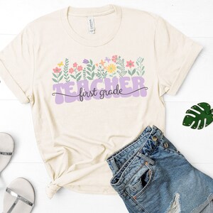 May include: A beige t-shirt with a purple graphic that says "Teacher first grade" with colourful flowers and leaves around the text.