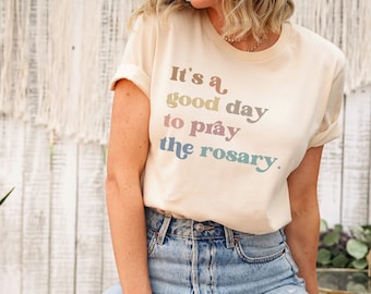 May include: A woman wearing a light beige t-shirt with the text "It's a good day to pray the rosary." in rainbow colors. She is wearing blue jeans with distressed details.