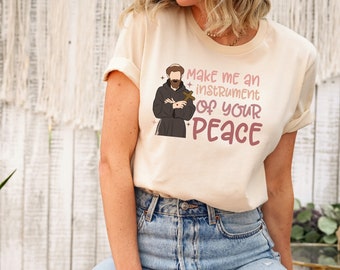 May include: A beige t-shirt featuring a graphic of a man holding a cross and the text "Make me an instrument of your peace".