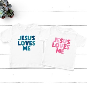 May include: Two white t-shirts with short sleeves. The left shirt has the text "JESUS LOVES ME" in blue and teal. The right shirt has the text "JESUS LOVES ME" in pink and red.