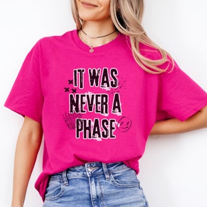 May include: A pink t-shirt with the text "It was never a phase" in black with a graphic design of a smiley face and x's.