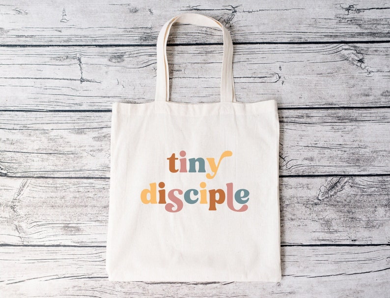 以下が含まれることがあります： カラフルなスタイリッシュな文字で「tiny disciple」と印刷された白いキャンバス地のトートバッグです。