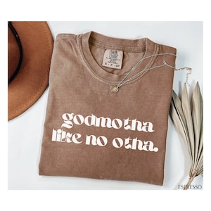 May include: A brown t-shirt with the text "godmotha like no otha." printed in white. The shirt is folded and laid on a white surface.