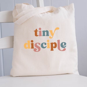 May include: A white canvas tote bag with the text "tiny disciple" printed in colorful letters. The letters are in shades of orange, yellow, brown, blue, and pink.