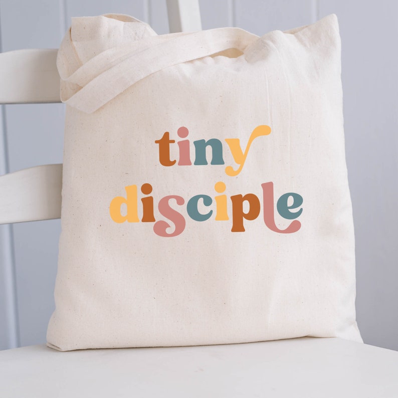 May include: A white canvas tote bag with the text "tiny disciple" printed in colorful letters. The letters are in shades of orange, yellow, brown, blue, and pink.