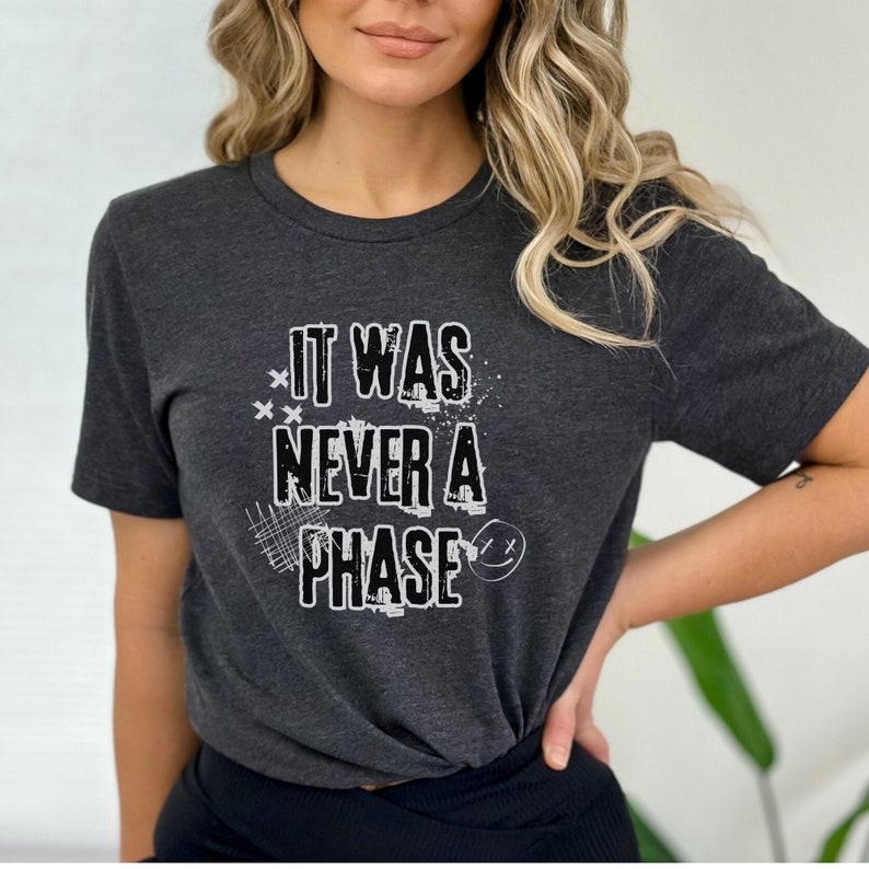 Może przedstawiać: Szary t-shirt z białym nadrukiem graficznym, na kt&oacute;rym widnieje napis "It Was Never A Phase" z uśmiechniętą buźką.