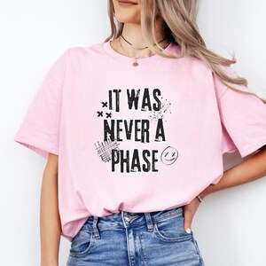 Może przedstawiać: R&oacute;żowa koszulka z czarnym napisem "It was never a phase" i uśmiechniętą buźką.