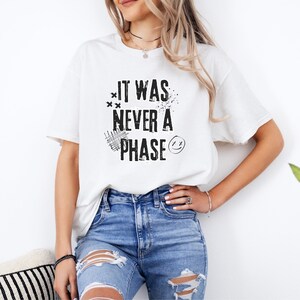 Może przedstawiać: Biała koszulka z czarnym napisem "It was never a phase" i uśmiechniętą buźką.