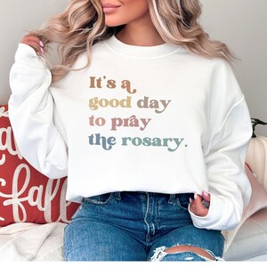 May include: White sweatshirt with the text "It's a good day to pray the rosary." in rainbow colors.