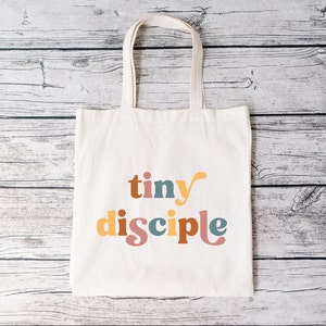 May include: A white canvas tote bag with the words "tiny disciple" printed in colorful letters. The letters are in a retro font style and are in shades of yellow, orange, pink, blue, and brown.