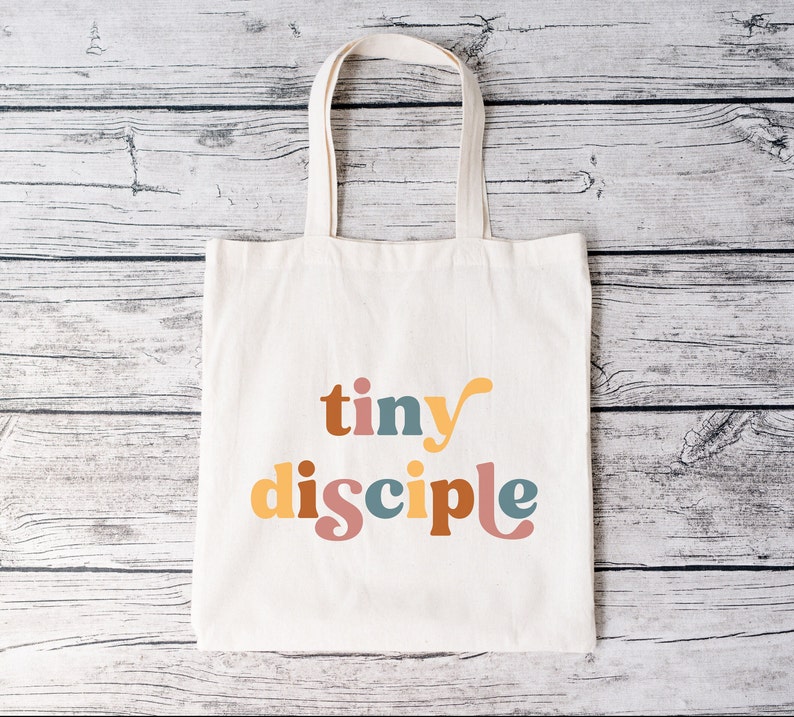 May include: A white canvas tote bag with the words "tiny disciple" printed in colorful letters. The letters are in a retro font style and are in shades of yellow, orange, pink, blue, and brown.
