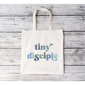 May include: A white canvas tote bag with the text "tiny disciple" in blue, green, and gray letters.