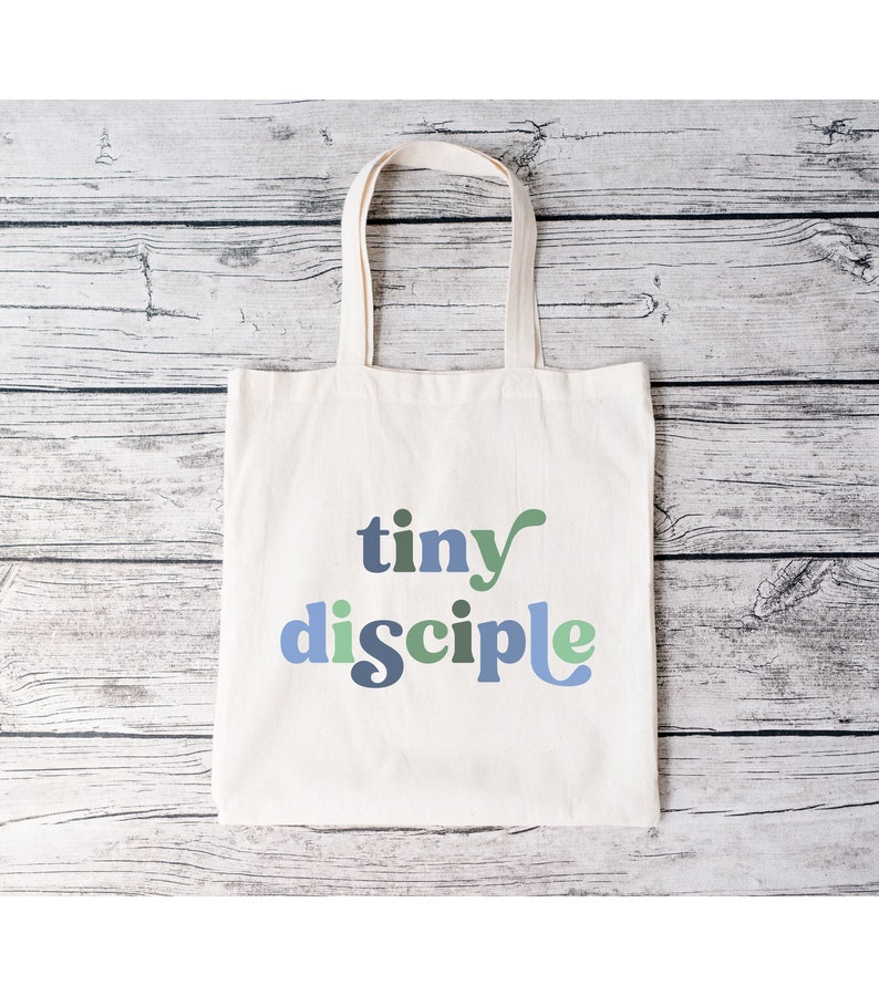 May include: A white canvas tote bag with the text "tiny disciple" in blue, green, and gray letters.
