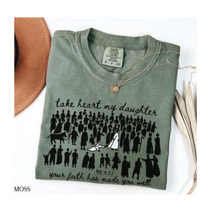 May include: Moss green Comfort Colors t-shirt with the text "take heart my daughter" and "your faith has made you well." The shirt features a graphic of people. A gold necklace and a brown hat are also visible.