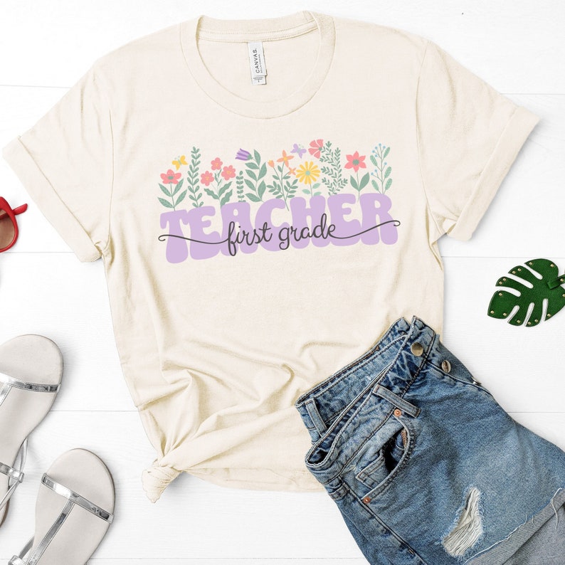 May include: A beige t-shirt with a purple graphic that says "Teacher first grade" with colourful flowers and leaves around the text.