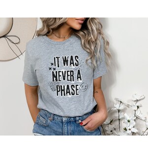 Może przedstawiać: Szary T-shirt z nadrukiem "It was never a phase" w kolorze czarnym, ze stylizowaną buźką i innymi elementami graficznymi.