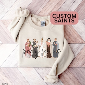 May include: A beige sweatshirt with a graphic print of a group of saints with the text "pray for us". The text "CUSTOM SAINTS" is in a pink box at the top of the image.