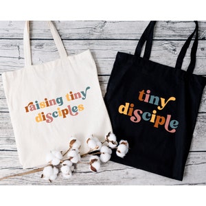以下が含まれることがあります： 異なるデザインのトートバッグ2つ。1つは白地に「raising tiny disciples」というカラフルな文字が書かれたバッグ。もう1つは黒地に「tiny disciple」というカラフルな文字が書かれたバッグです。