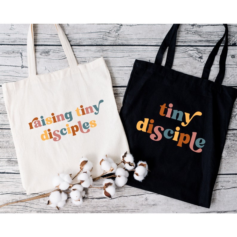 以下が含まれることがあります： 異なるデザインのトートバッグ2つ。1つは白地に「raising tiny disciples」というカラフルな文字が書かれたバッグ。もう1つは黒地に「tiny disciple」というカラフルな文字が書かれたバッグです。