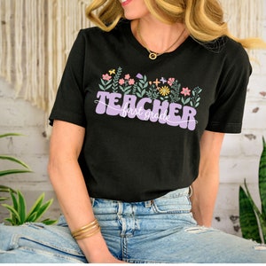 May include: A black t-shirt with a purple "Teacher first grade" graphic featuring colourful flowers and leaves.