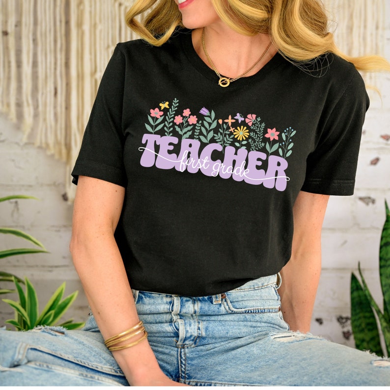 May include: A black t-shirt with a purple "Teacher first grade" graphic featuring colourful flowers and leaves.